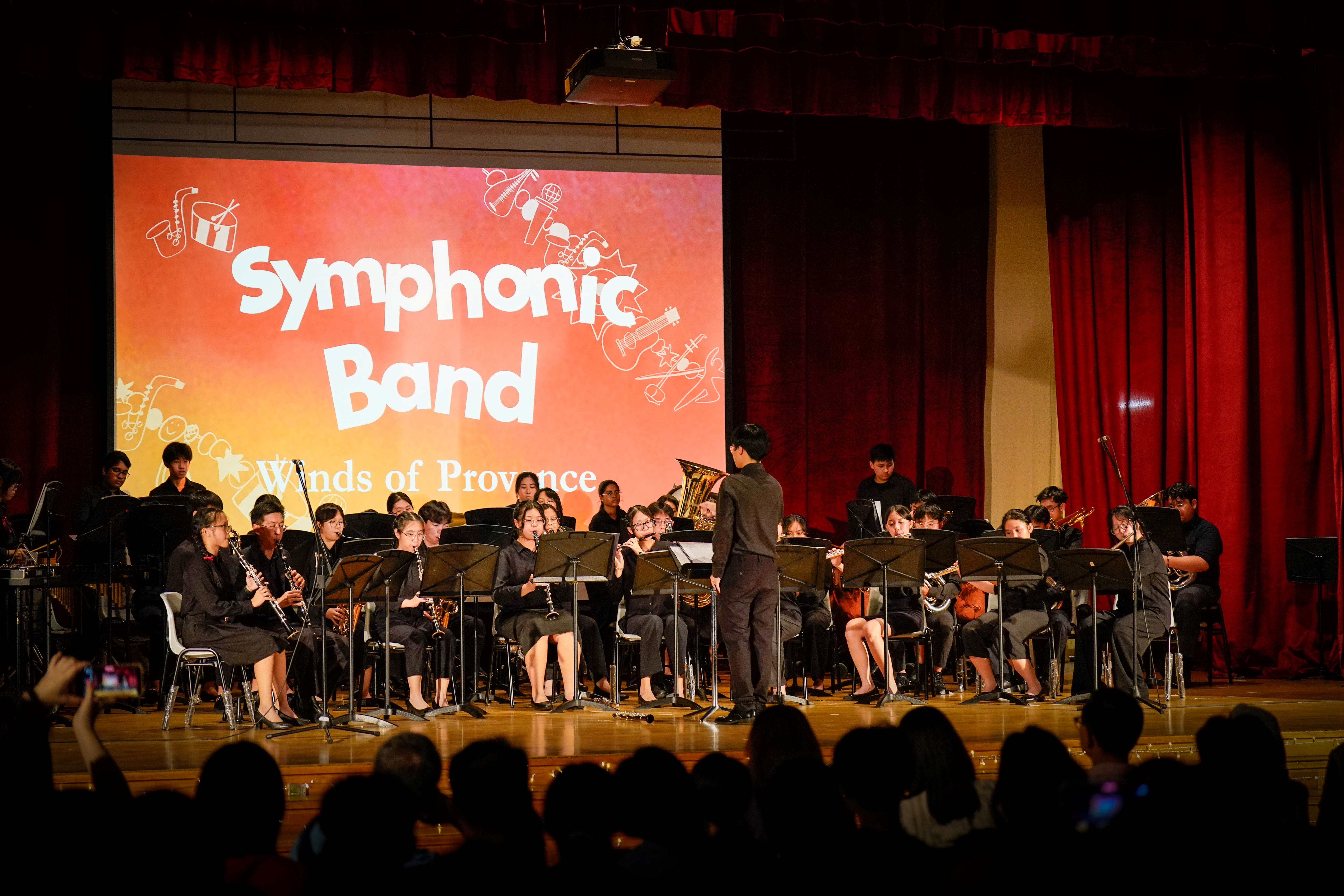 A Symphonic Band performs "Winds of Provence" on stage, with students in black playing wind and brass instruments under a conductor.